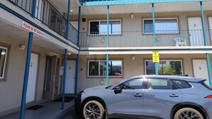 Un motel de dos pisos con fachada blanca y detalles azules. Hay un letrero que indica una política de 'No Invertir el Vehículo'. Un coche está estacionado al frente, y un letrero de precaución advierte sobre pisos resbaladizos cuando están mojados.