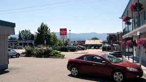 Un estacionamiento con un coche rojo aparcado en primer plano, un edificio con el rótulo de Boston Pizza, y montañas al fondo.