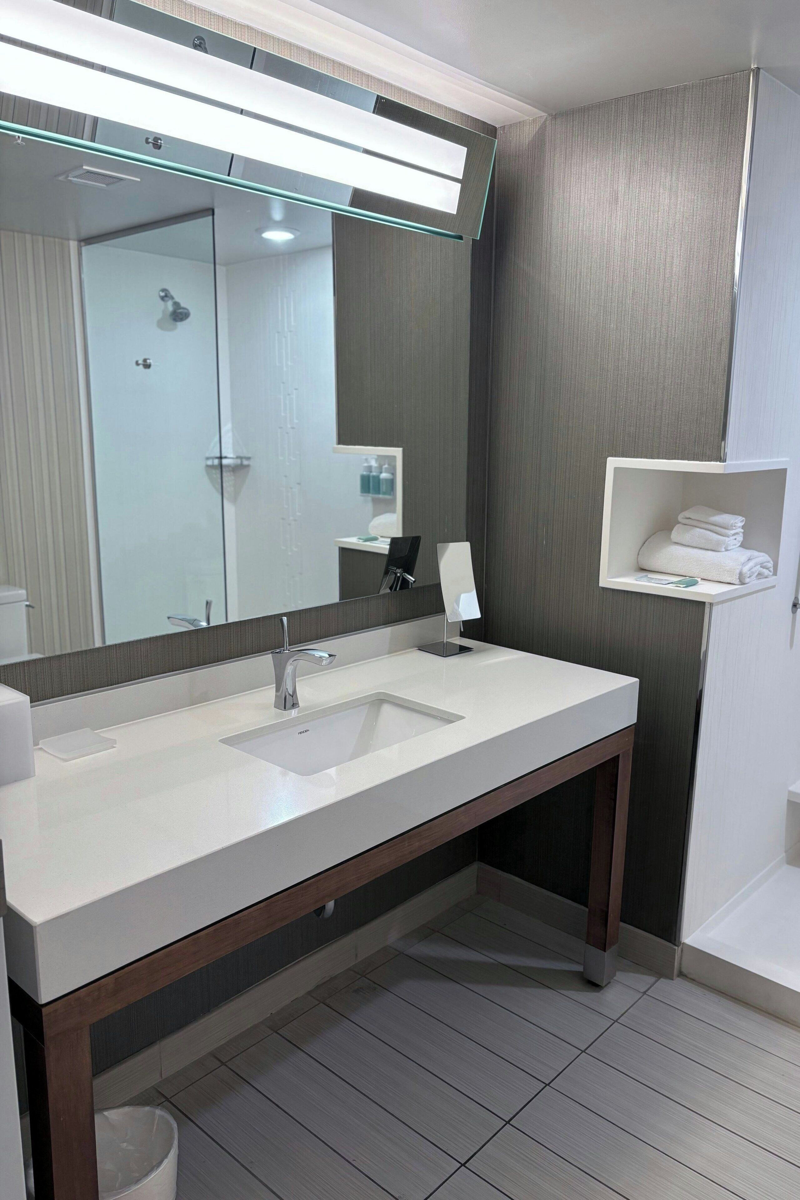 A modern bathroom with a large mirror, two sinks, a vanity, and a towel shelf.