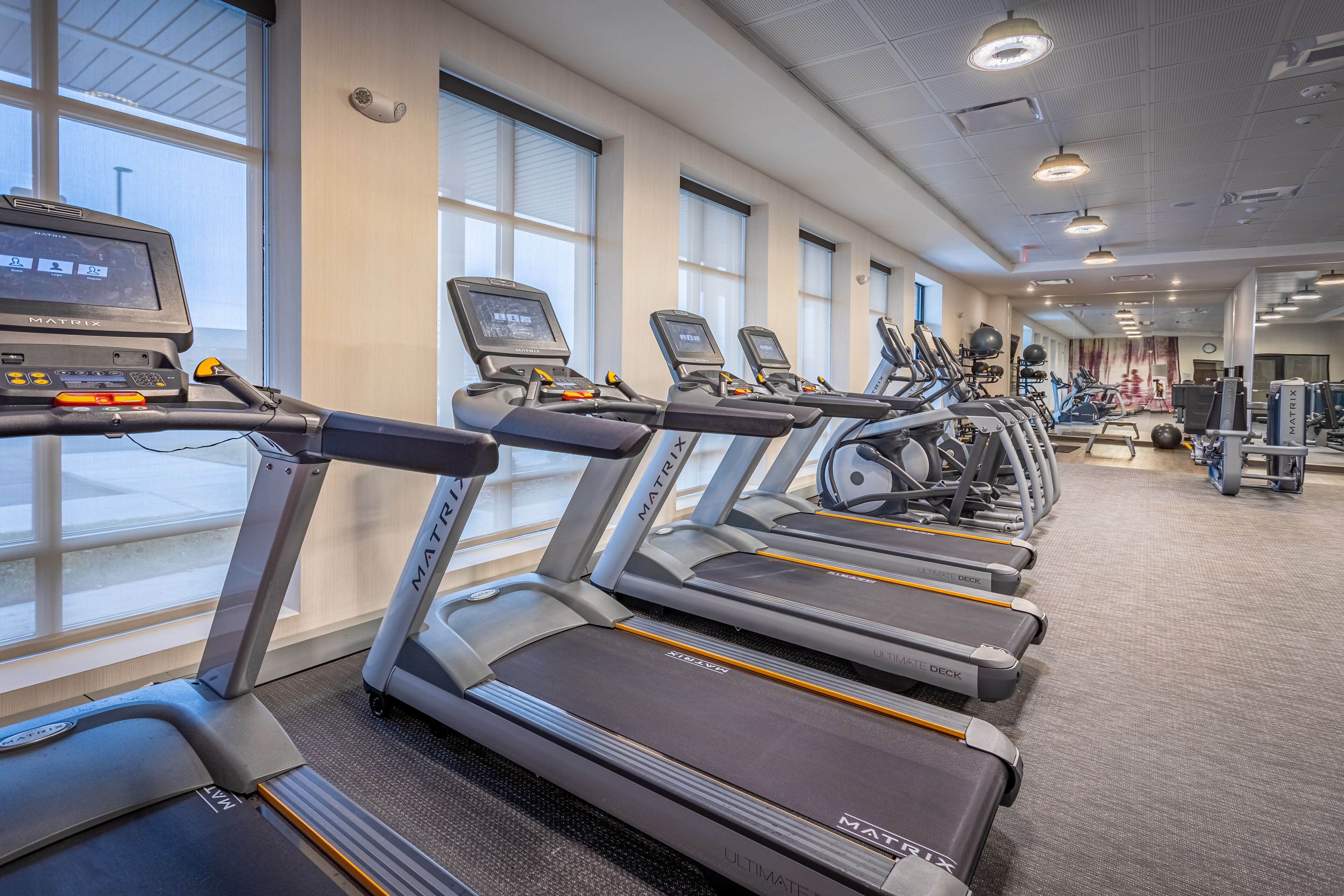 A row of treadmills with digital displays, arranged in a gym with large windows and various exercise equipment.