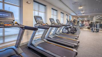 A row of treadmills with digital displays, arranged in a gym with large windows and various exercise equipment.