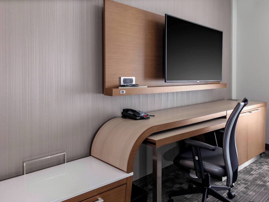 A hotel room with a wooden desk, a flat-screen TV mounted on the wall, a telephone, and a chair.