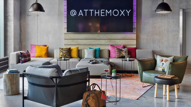 Ett modernt vardagsrum med en hörnsoffa, en grön fåtölj och ett soffbord. En neonskylt med texten "@atthemoxy" hänger på väggen.