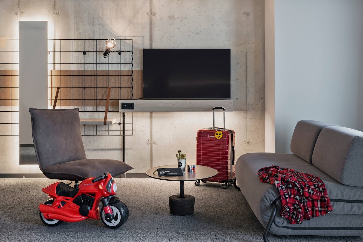 A modern living room with a large flat-screen TV mounted on the wall, a red suitcase, a grey sofa, a brown chair, and a small round table with a book and a Rubik's cube.
