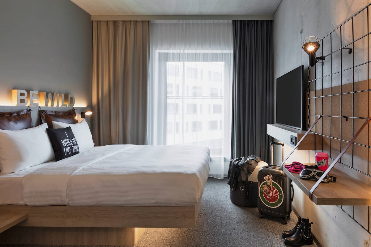 A hotel room with a bed, a TV mounted on the wall, a suitcase, and a pair of boots.