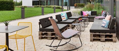 An outdoor seating area with modern furniture, including a rocking chair and a sofa, set against a backdrop of a contemporary building with large windows.
