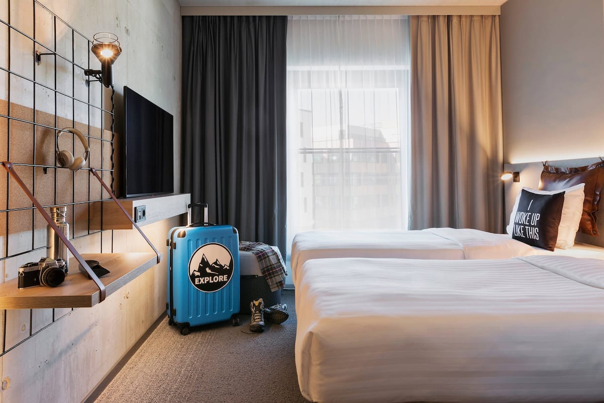 A hotel room with a bed, a desk with a lamp, a suitcase, and a television mounted on the wall.