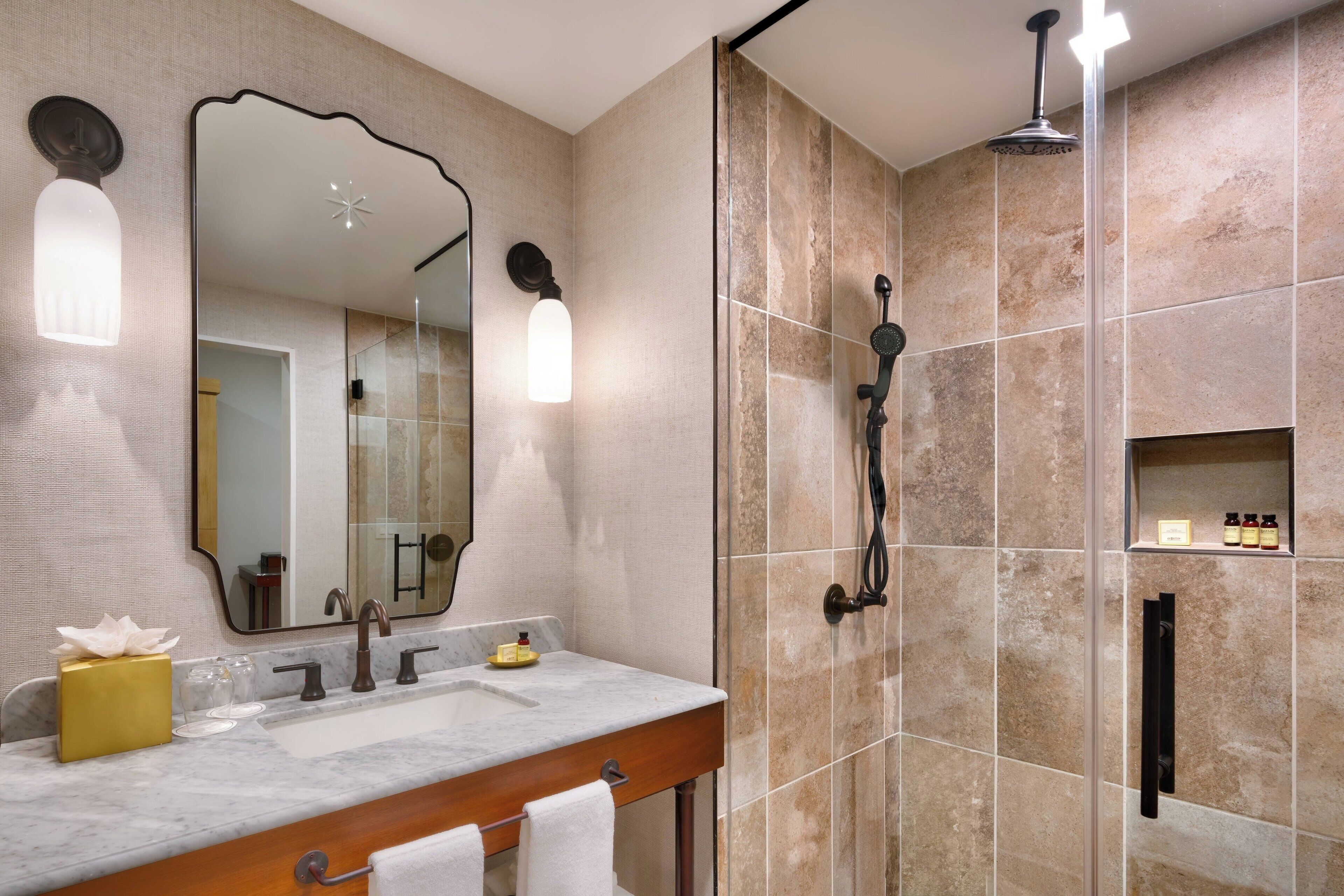Une salle de bain avec un lavabo en marbre, un grand miroir, une douche avec un pommeau de douche amovible et une étagère contenant des articles de toilette.