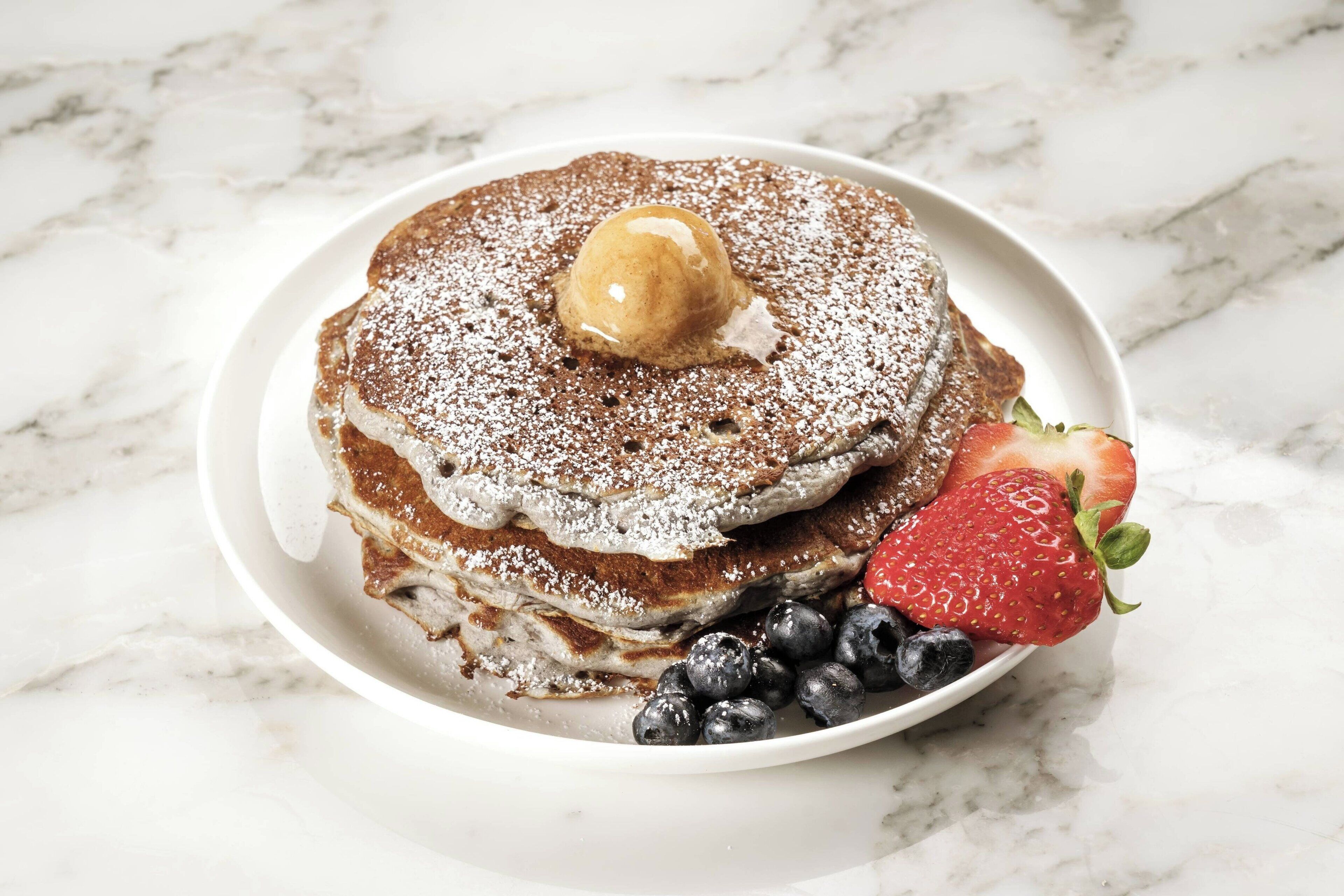 A stack of pancakes with a dollop of cream and powdered sugar on top, accompanied by a strawberry and blueberries.
