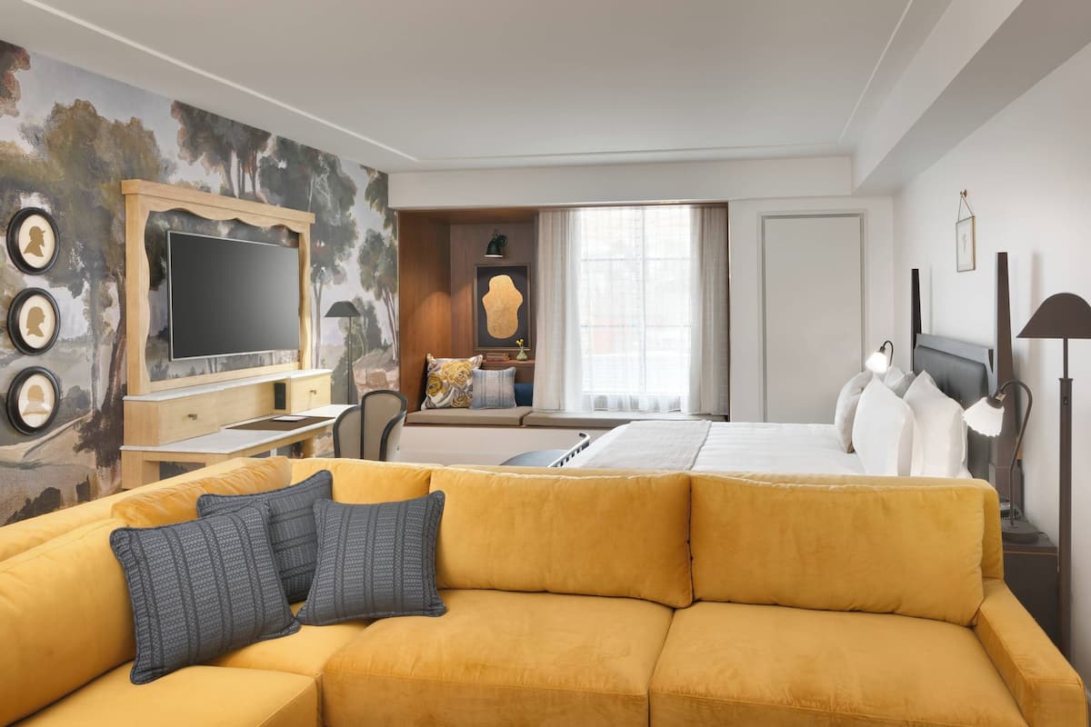 Ein Hotelzimmer mit gelber Couch, einem Bett, einem Fernseher, einem Schreibtisch und einem Fenster mit Vorhängen.