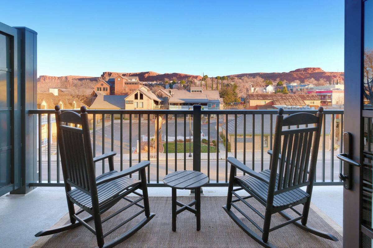Ein Balkon mit zwei Schaukelstühlen und einem kleinen Tisch, mit Blick auf eine Stadt mit Gebäuden und Hügeln im Hintergrund.