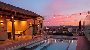 A rooftop pool with a view of a church spire, string lights, and a covered seating area.