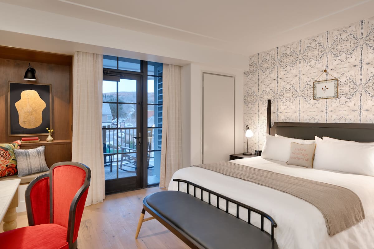 Ein Hotelzimmer mit einem großen Bett, einem Schreibtisch mit Stuhl, einer Bank, einem Nachttisch mit Lampe und Blick ins Freie durch eine Schiebetür.