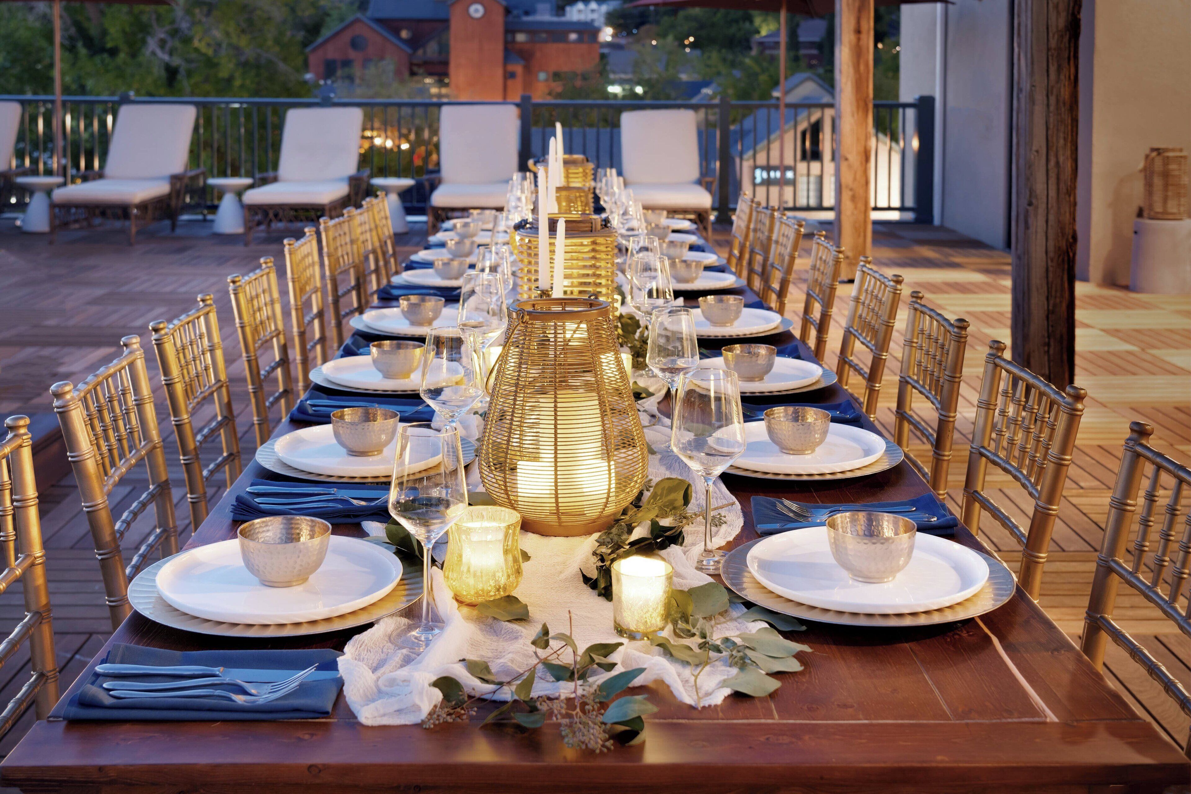 A long dining table set with white plates, gold-rimmed glasses, and candles, surrounded by bamboo chairs on an outdoor deck.