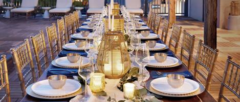 A long dining table set with white plates, gold-rimmed glasses, and candles, surrounded by bamboo chairs on an outdoor deck.
