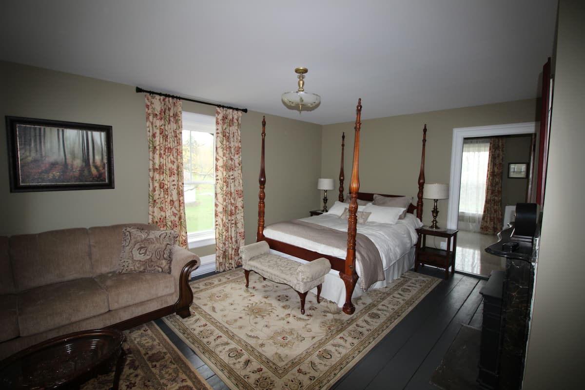 A four-poster bed with a wooden headboard, a sofa, a bench, and a patterned rug in a room with a large window and a painting on the wall.