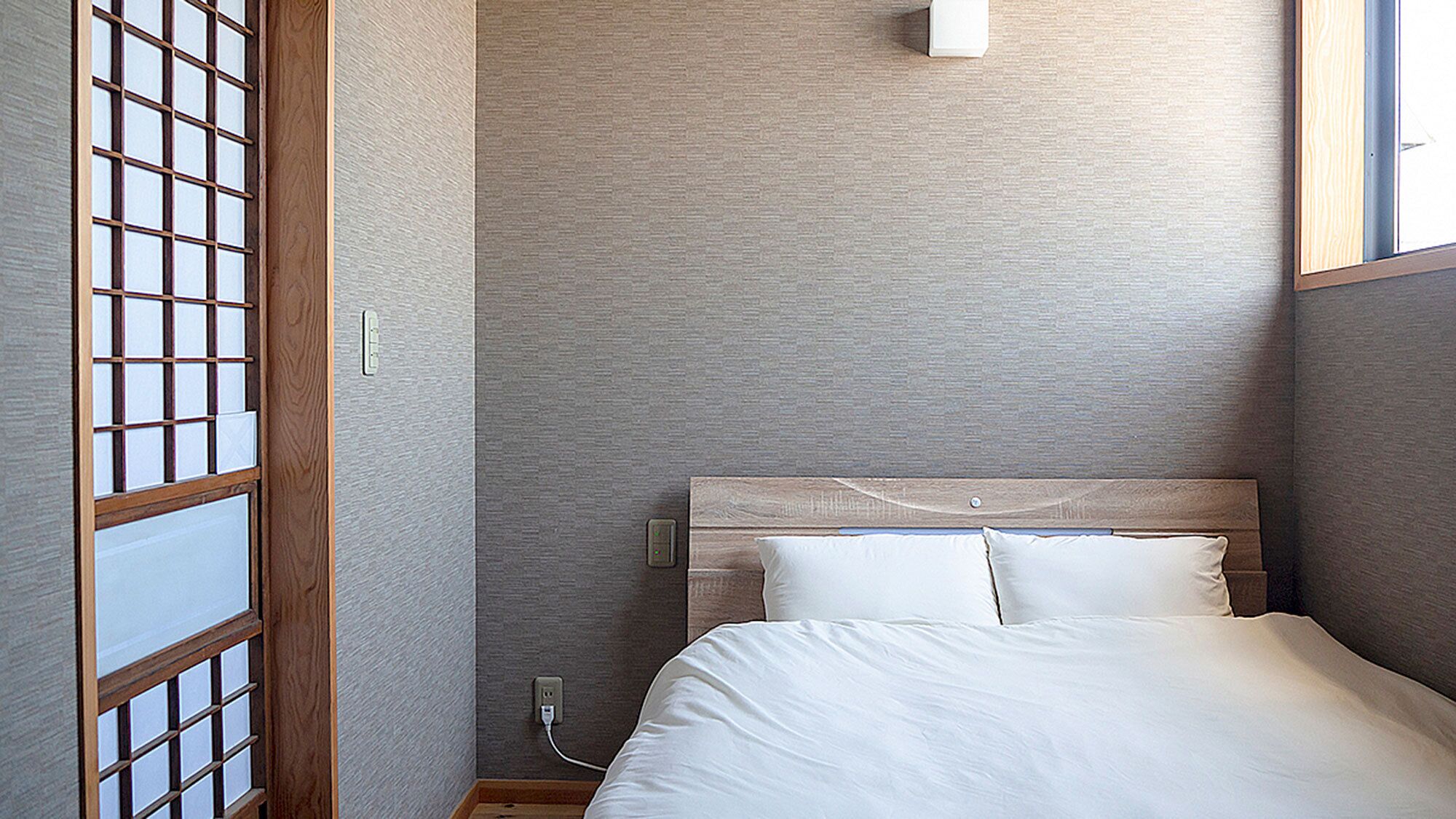 A room with a bed, a wooden headboard, a wall-mounted light, and a window with a view.
