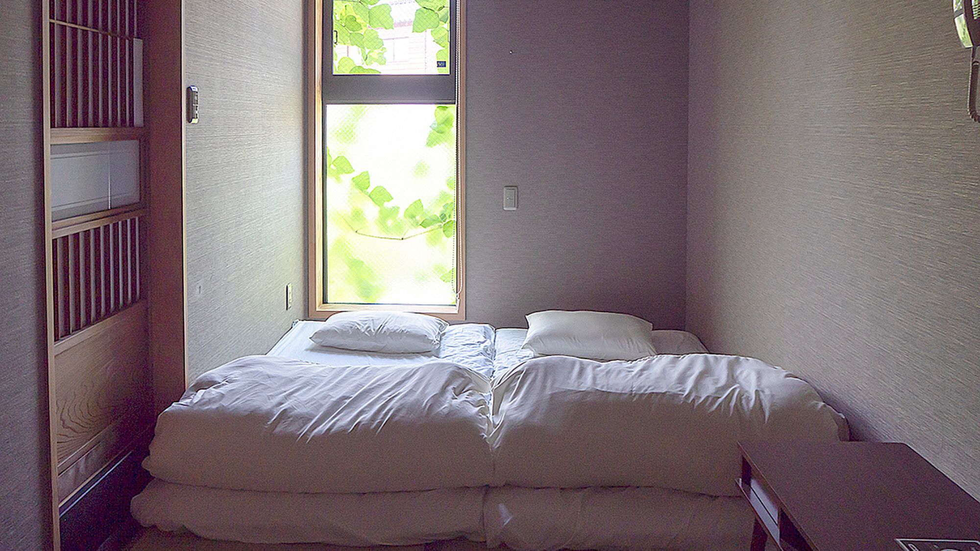 A small, neatly made bed in a room with a window, a radiator, and a wall-mounted light switch.
