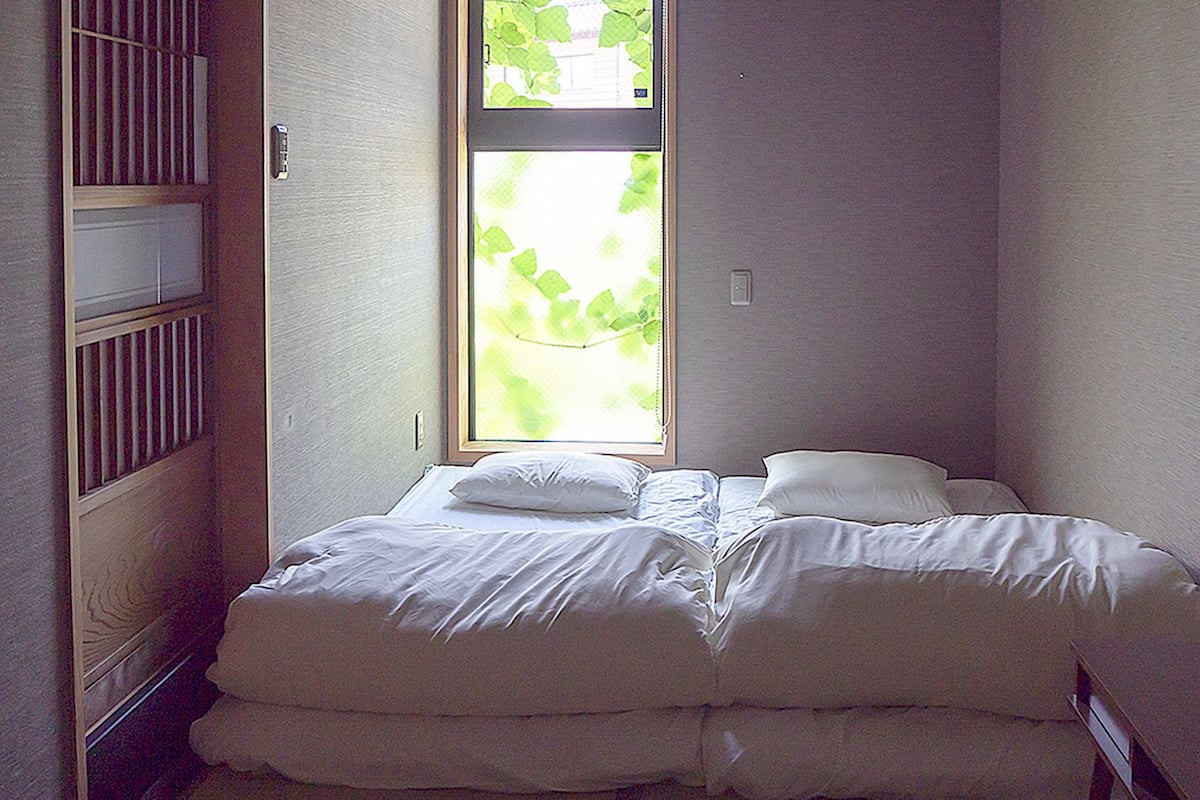 A small, neatly made bed in a room with a window, a radiator, and a wall-mounted light switch.