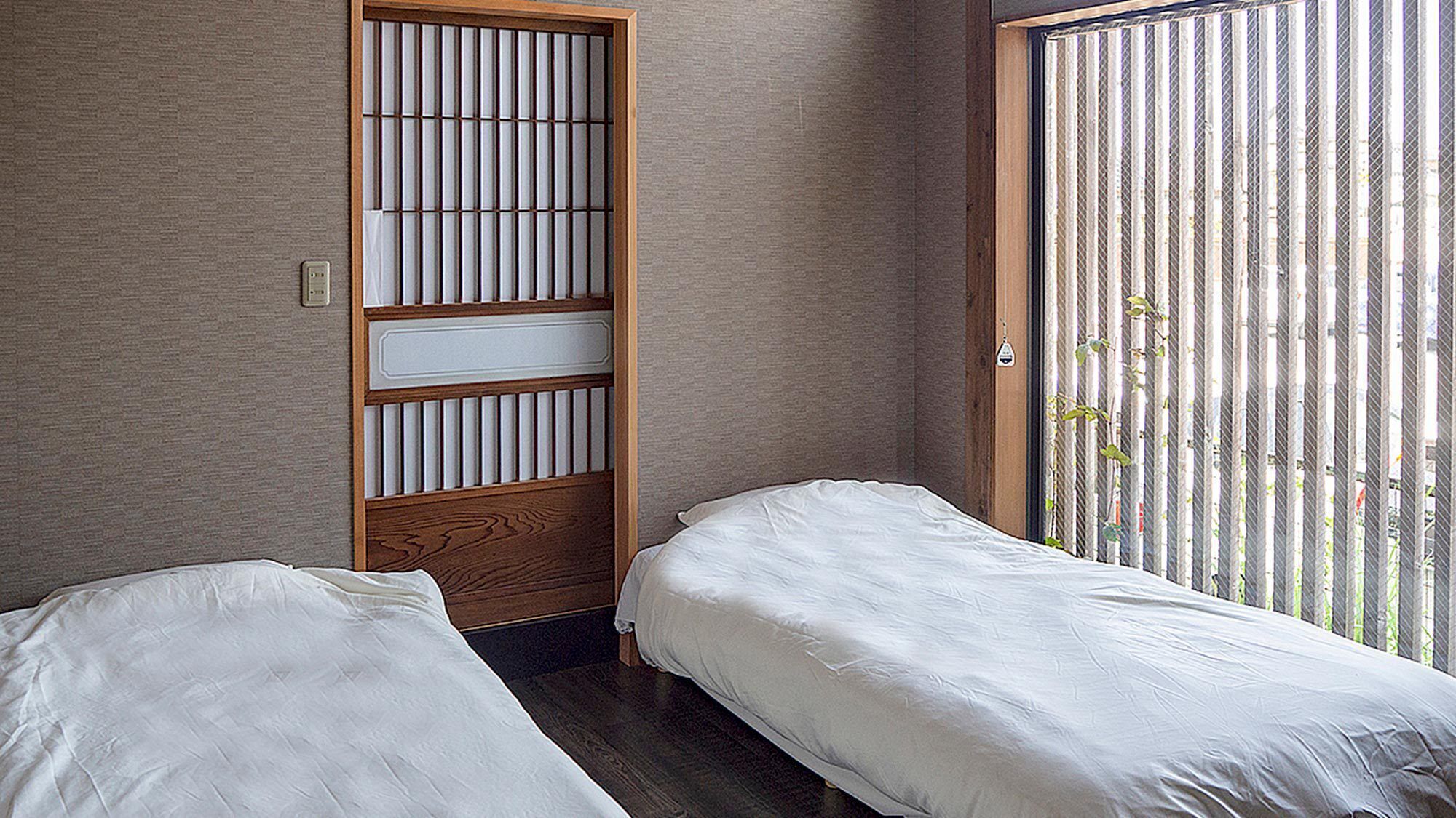 A room with two single beds, a wooden partition with vertical slats, and a window with vertical blinds.