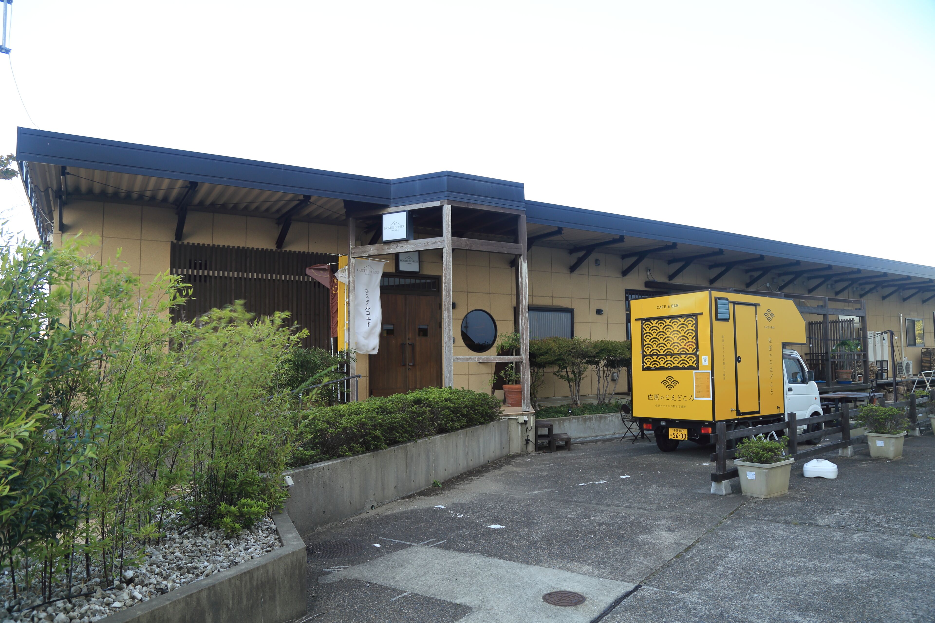 A building with a covered entrance, a yellow delivery truck parked in front, and potted plants along the sidewalk.