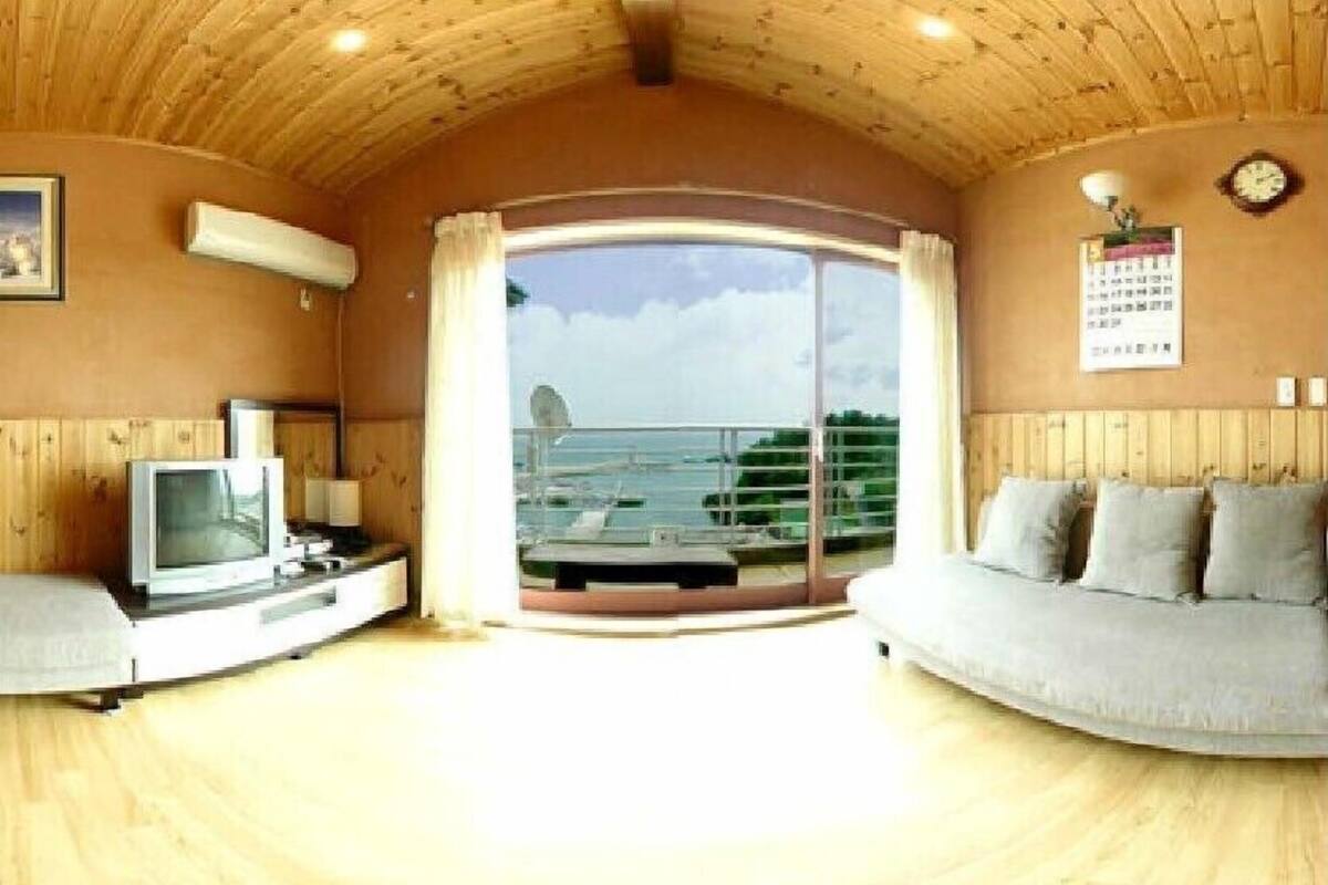 A room with a wooden ceiling, a television, a sofa, and a large window with a view of the sea.