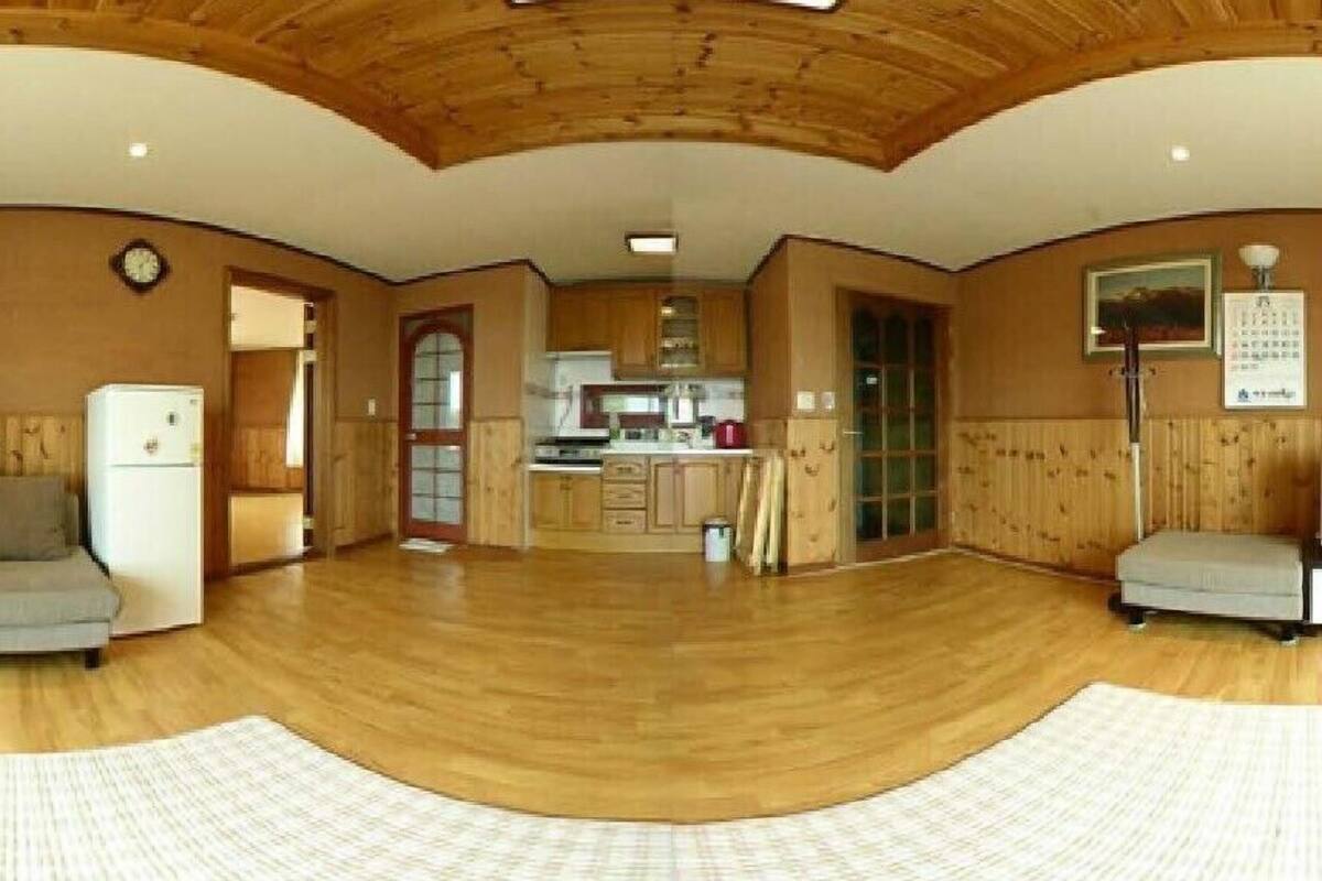 A spacious living area with wooden flooring, a kitchen in the background, and a large window.