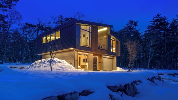 Una casa moderna de dos pisos con ventanas amplias y un garaje, rodeada de nieve y árboles.
