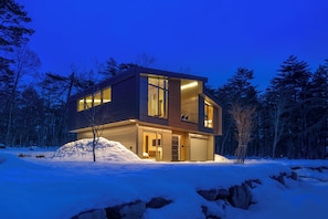 A modern two-story house with large windows and a garage, surrounded by snow and trees.