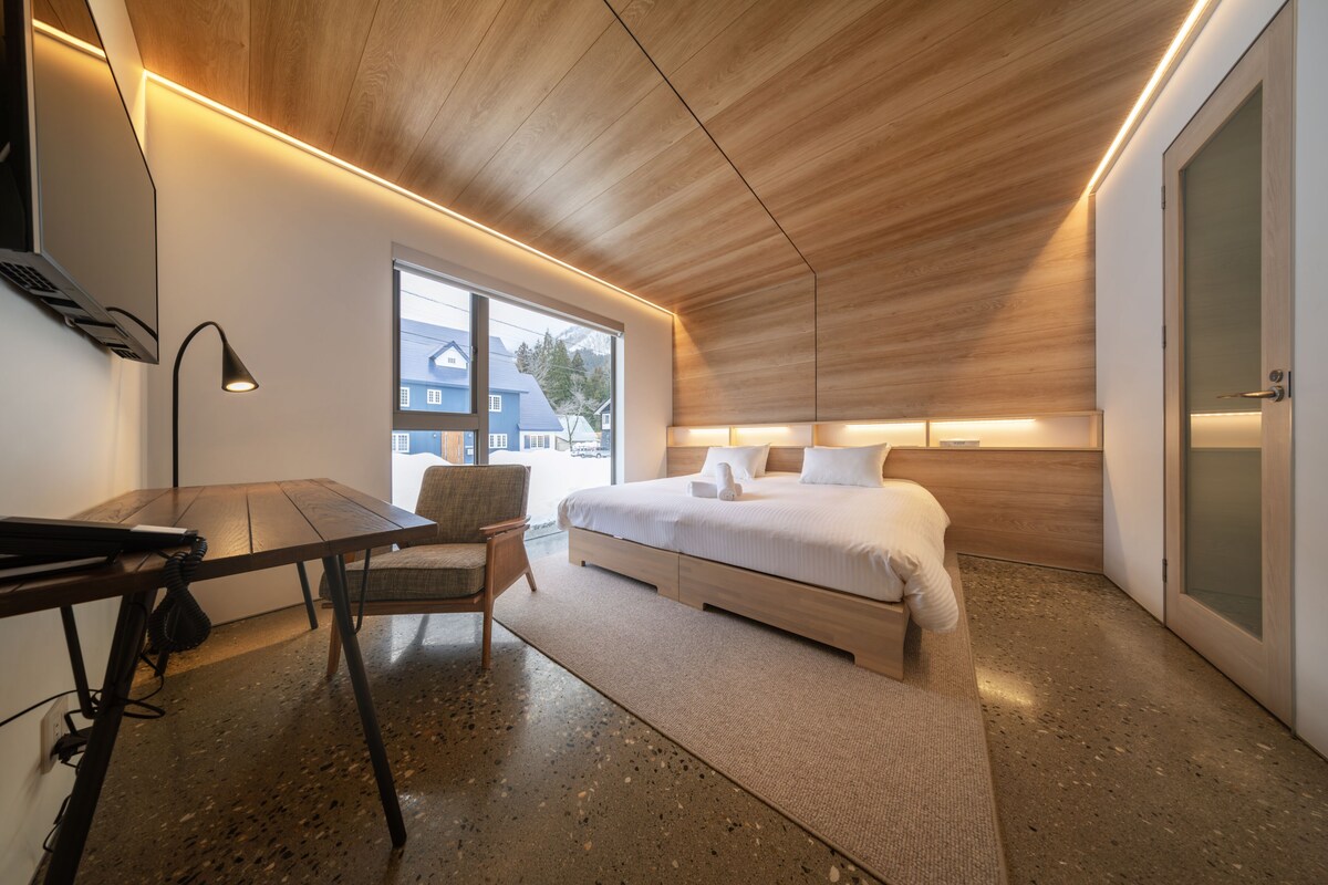 A modern bedroom with a wooden ceiling, a bed with white bedding, a desk with a chair, and a large window offering a view of buildings outside.