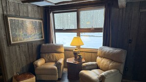 Um quarto acolhedor com duas poltronas de couro, uma pequena mesa de madeira com uma luminária, e uma janela ampla com vista para uma paisagem nevada.