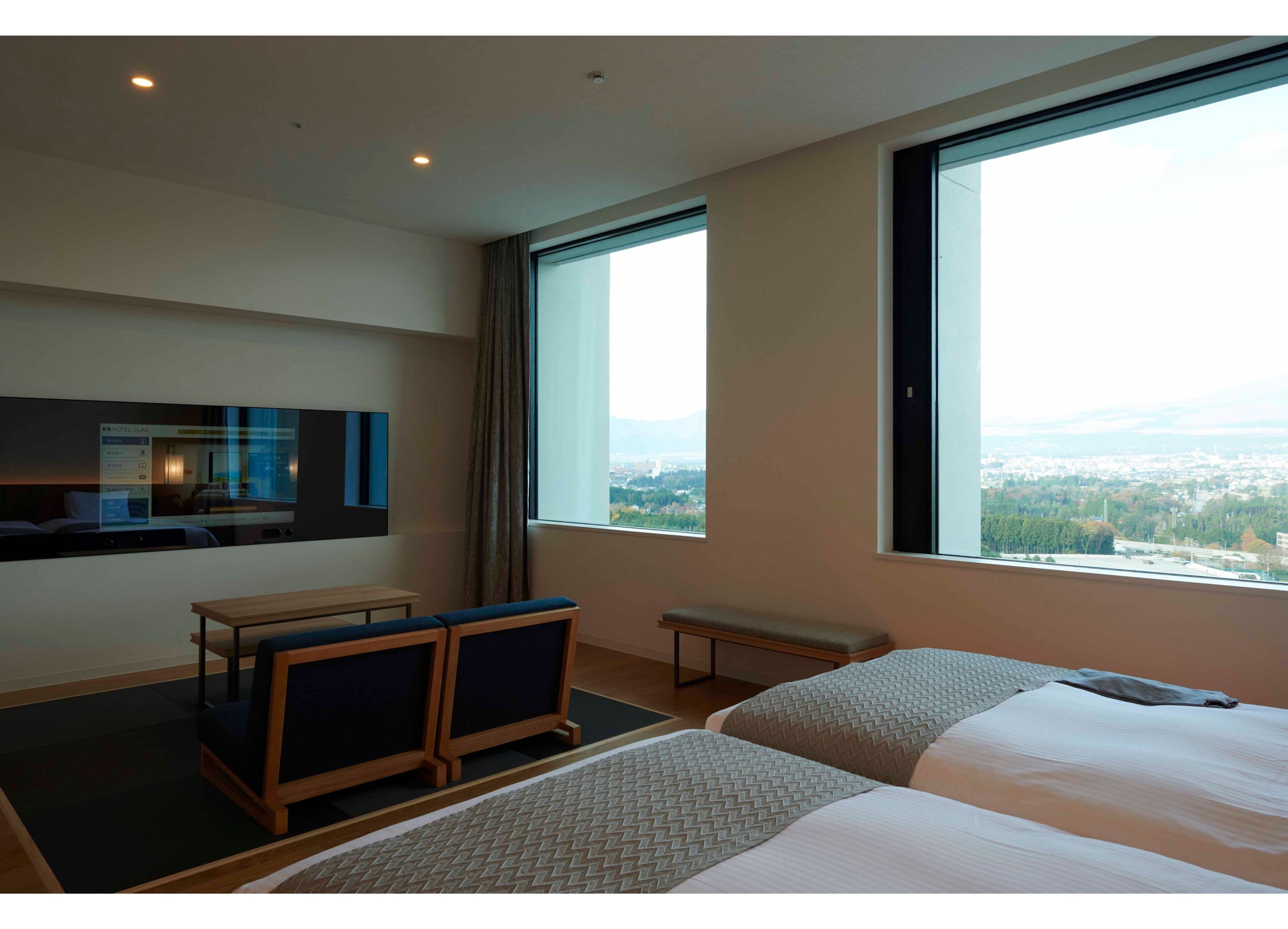 A hotel room with two beds, a desk, a chair, a TV, and large windows offering a city view.