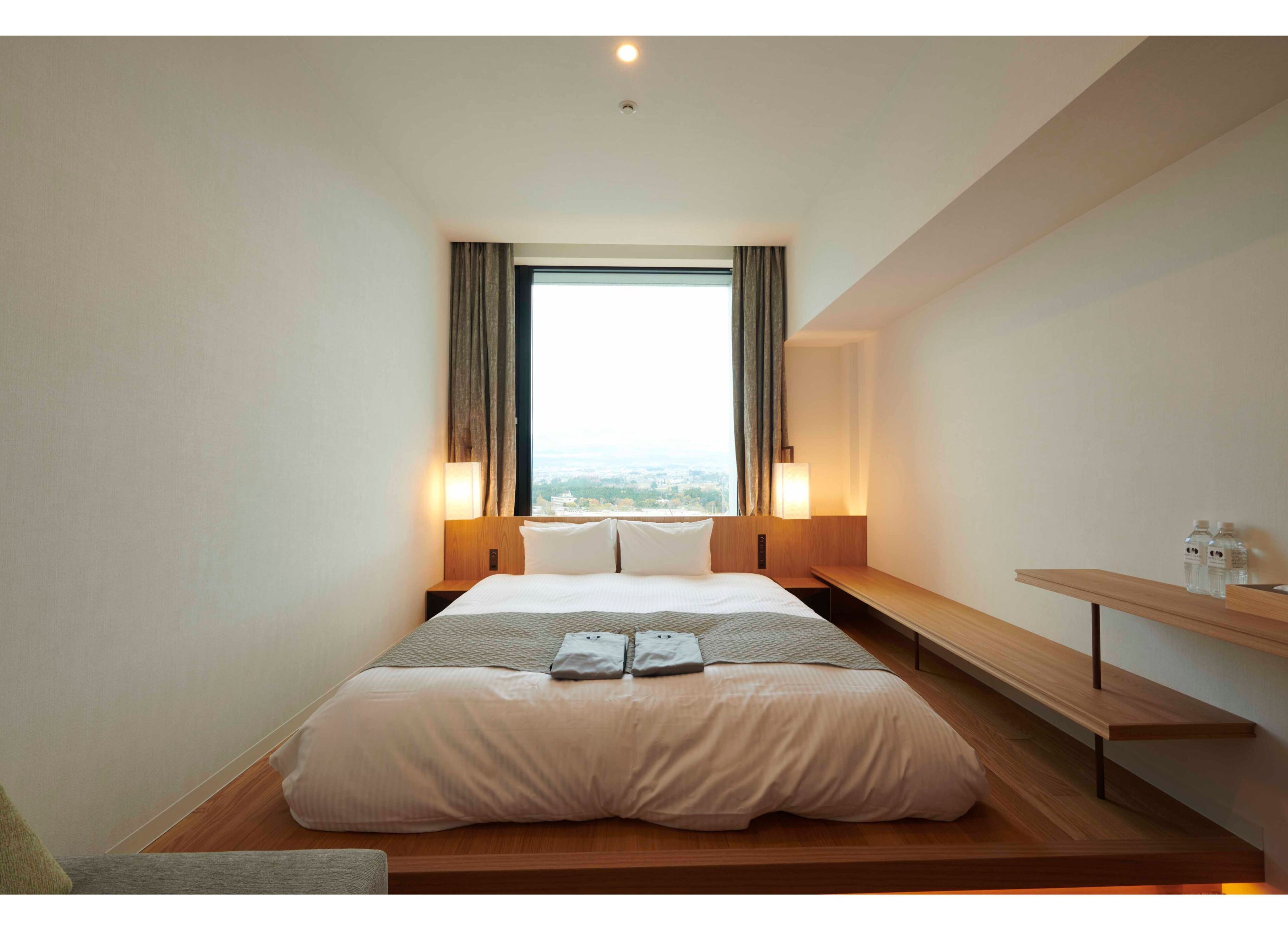 A modern hotel room with a large bed, bedside lamps, a bench, and a view of the city.
