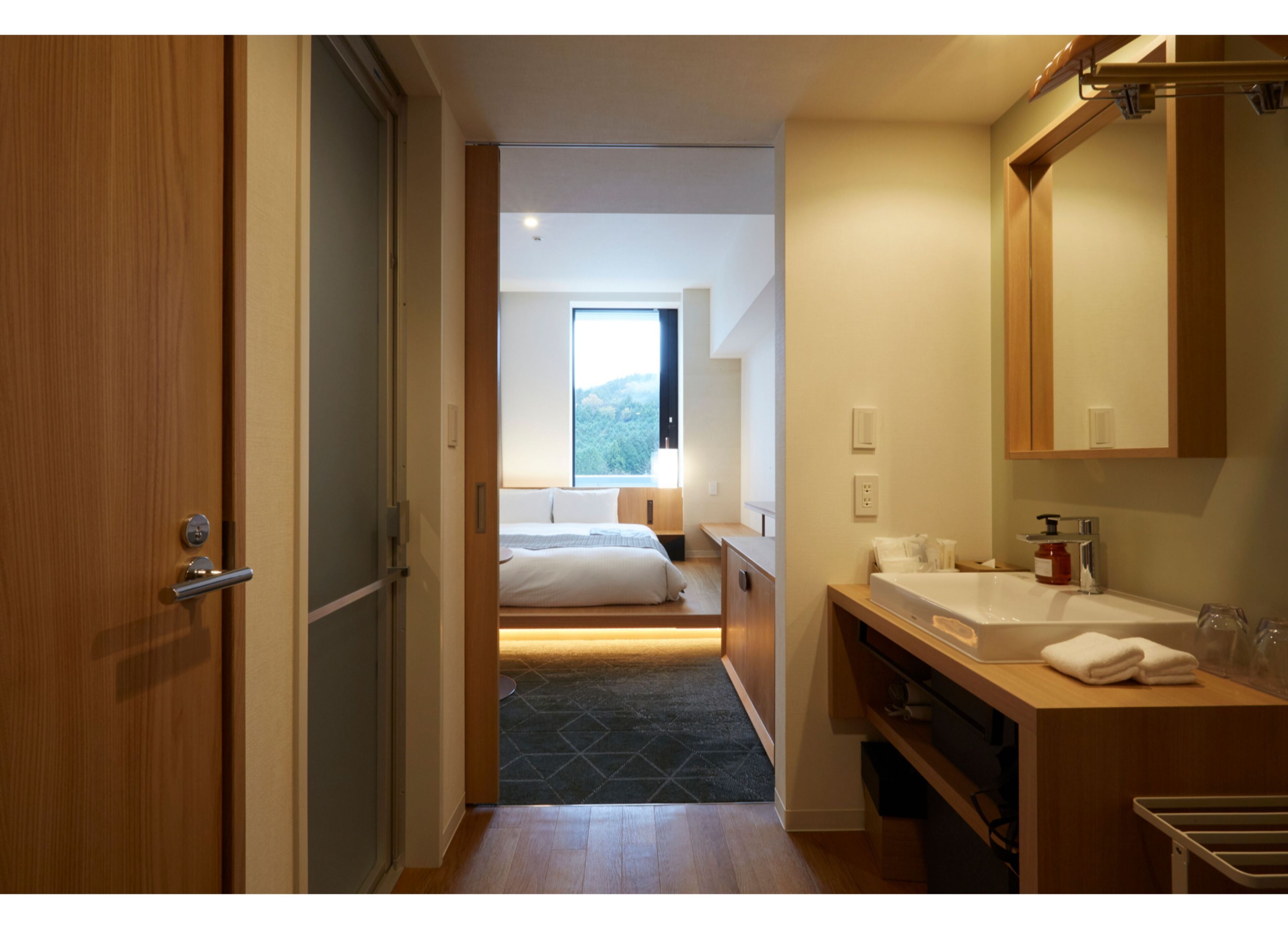 A hotel room with a wooden door, a bed with white linens, a bathroom with a sink and mirror, and a view of the outdoors through a window.