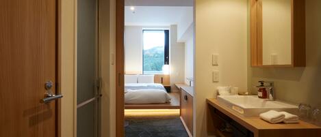 A hotel room with a wooden door, a bed with white linens, a bathroom with a sink and mirror, and a view of the outdoors through a window.