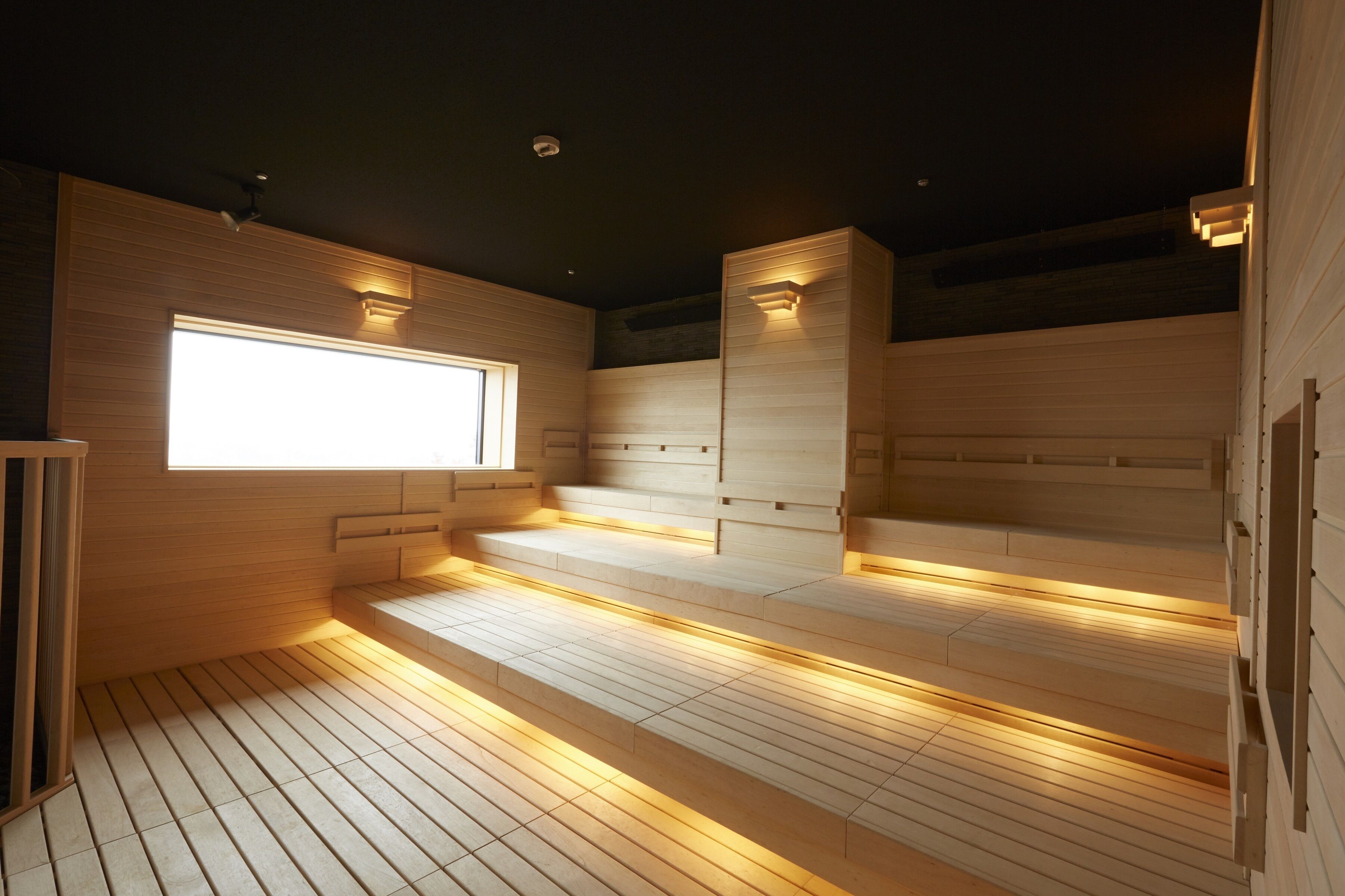 A sauna room with wooden benches and steps, a large window, and wall-mounted lighting.