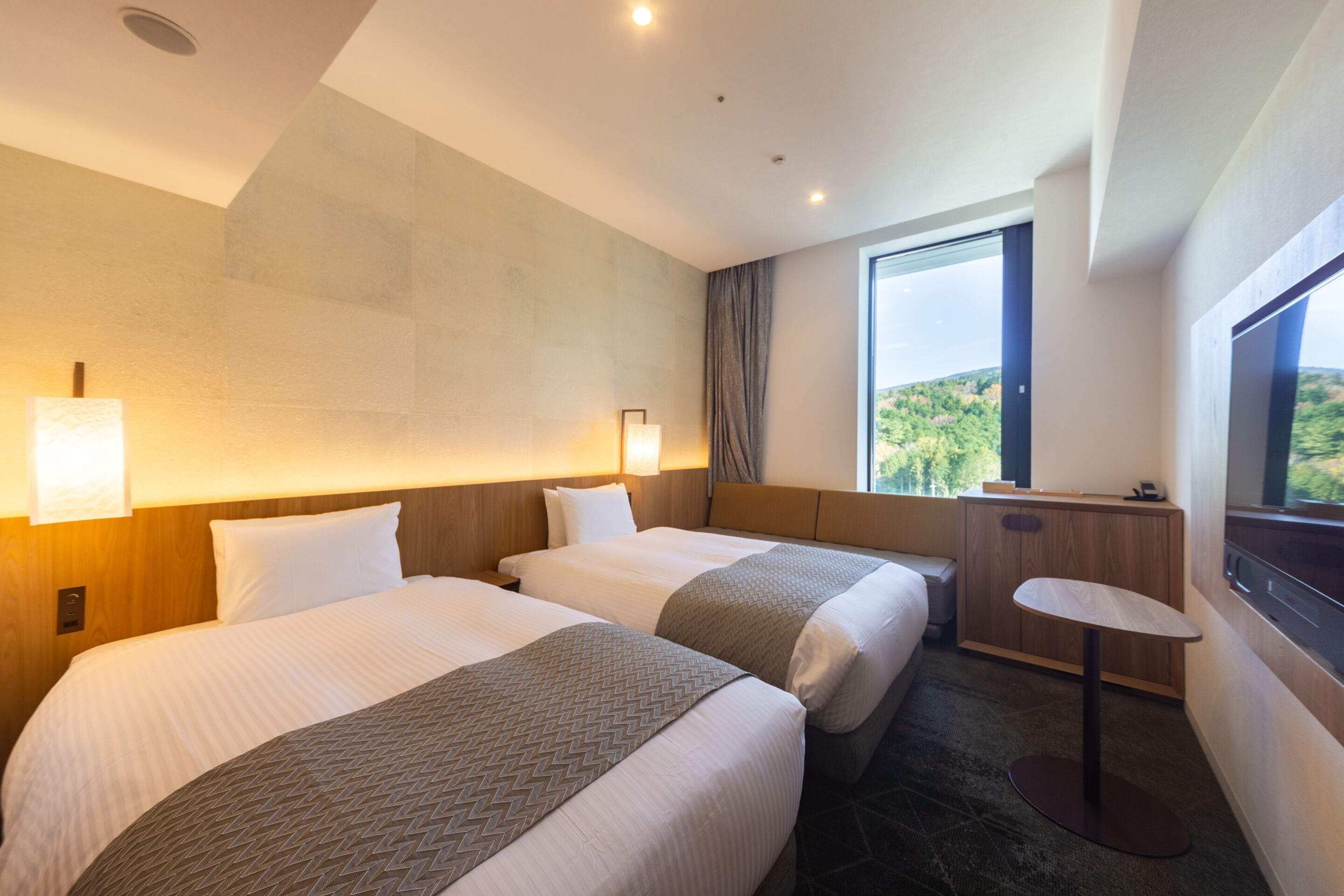 A hotel room with two beds, a small desk, a window with a view of greenery, and a ceiling with recessed lighting.