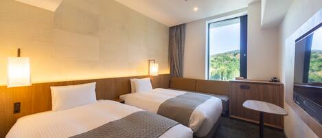 A hotel room with two beds, a small desk, a window with a view of greenery, and a ceiling with recessed lighting.