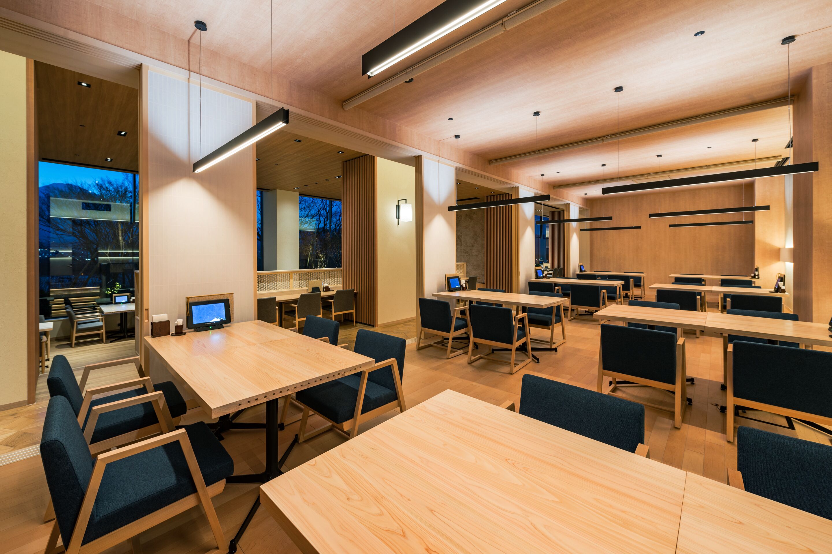 Una sala de conferencias moderna con varias mesas y sillas, suelo de madera y grandes ventanales que ofrecen vistas al exterior.