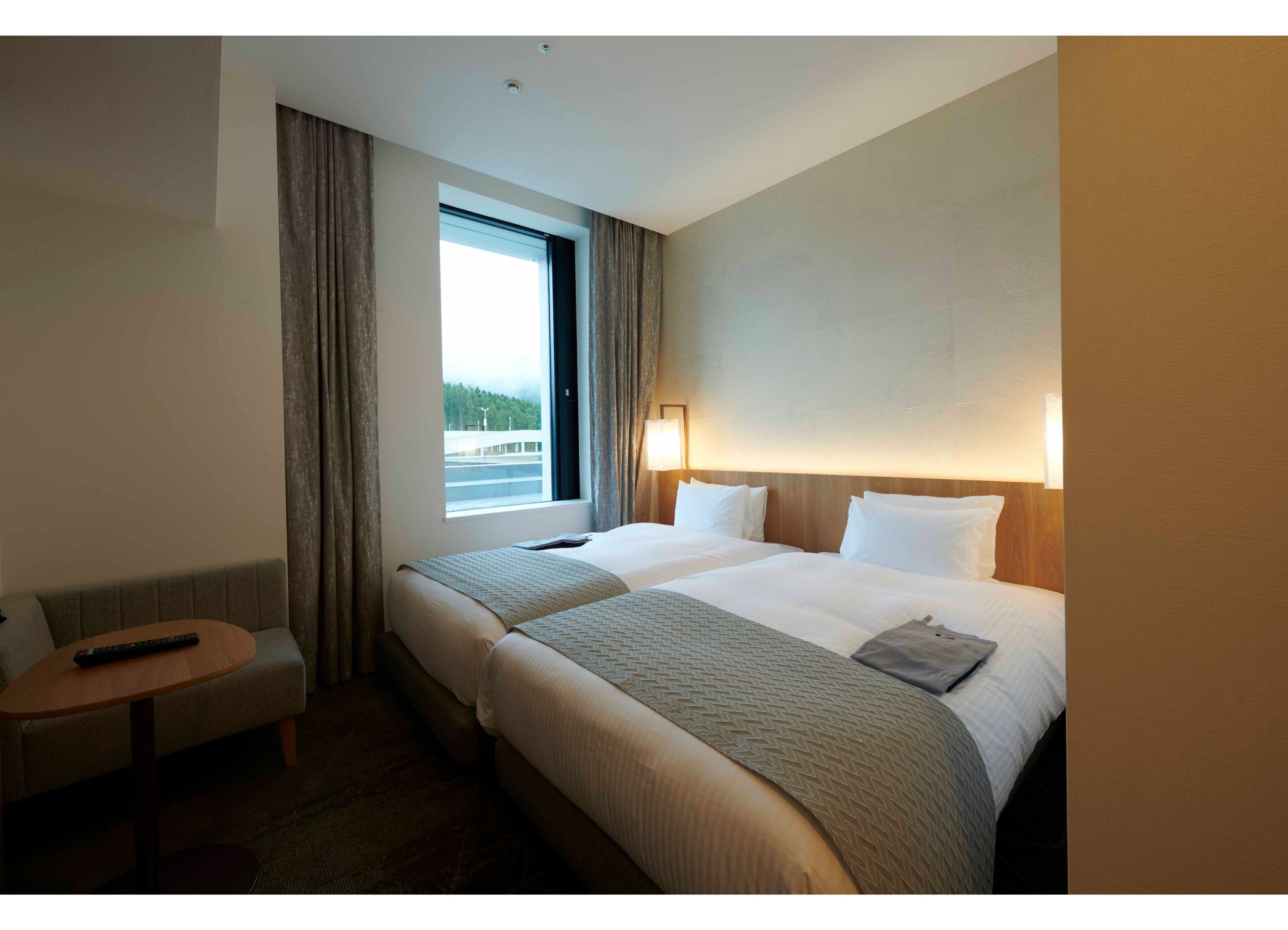 A hotel room with two beds, a small table, a remote control, and a window with a view of a bridge and greenery.