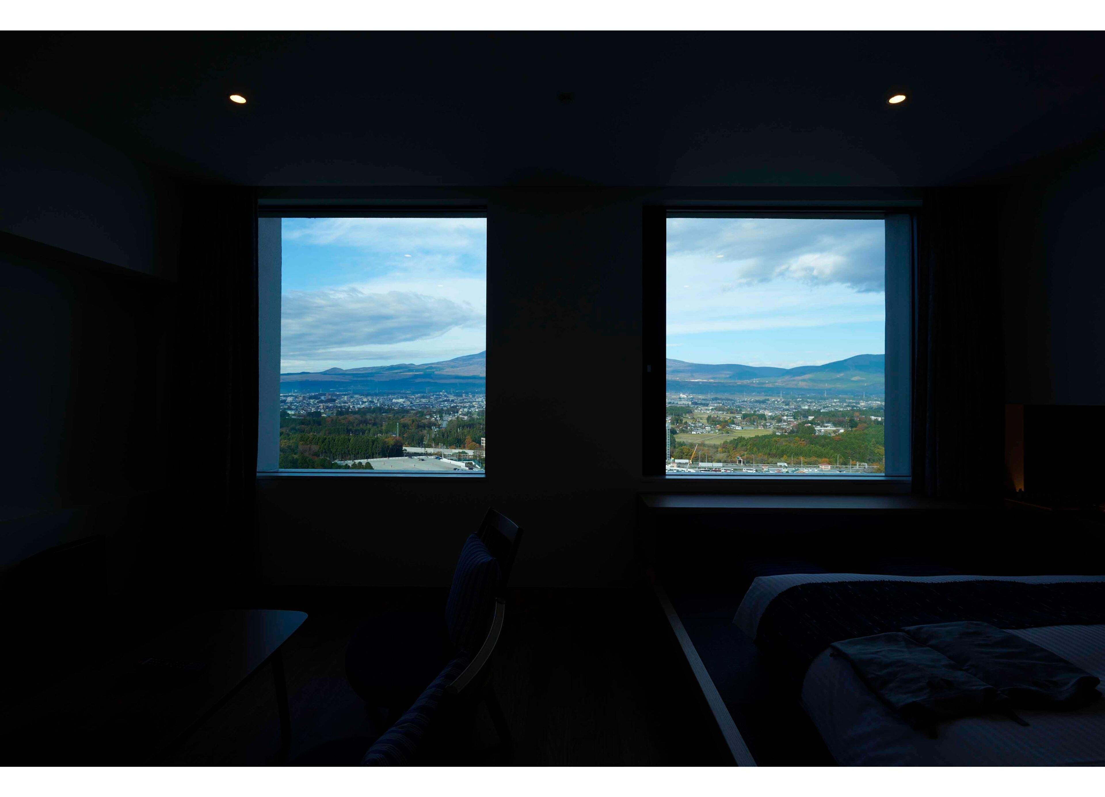 Une chambre d’hôtel dotée de deux grandes fenêtres donnant sur un paysage urbain et des montagnes.