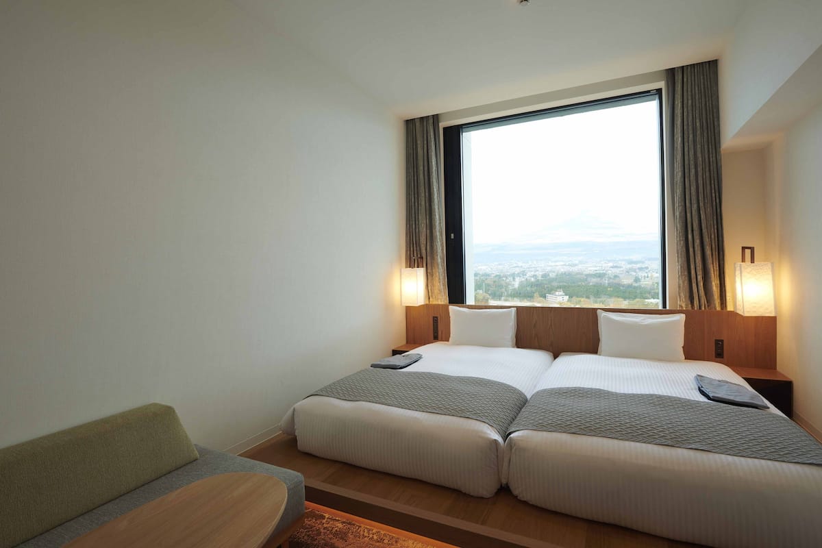 Ein Hotelzimmer mit zwei Betten, einem Sofa und Blick auf die Stadt, inklusive großem Fenster mit Vorhängen.