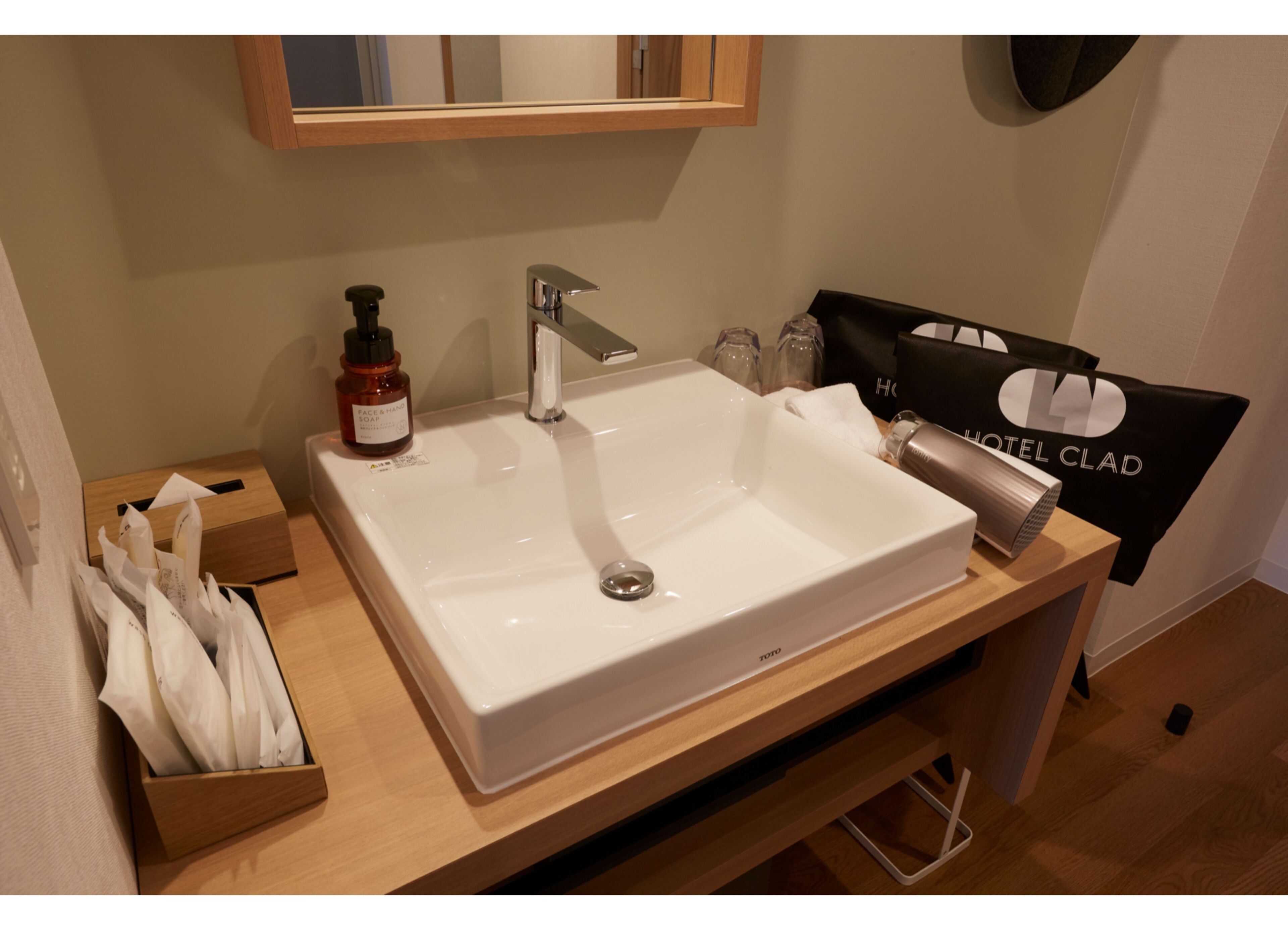 A hotel bathroom with a white sink, a mirror above it, a bottle of face wash, and a towel rack with a towel.