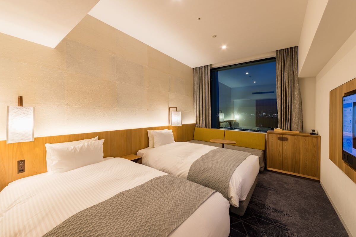 Ein Hotelzimmer mit zwei Betten, einem großen Fenster mit Stadtblick und einem an der Wand befestigten Fernseher.