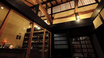 A bar area with wooden beams and a high ceiling, shelves stocked with bottles, and a large window with a grid pattern.