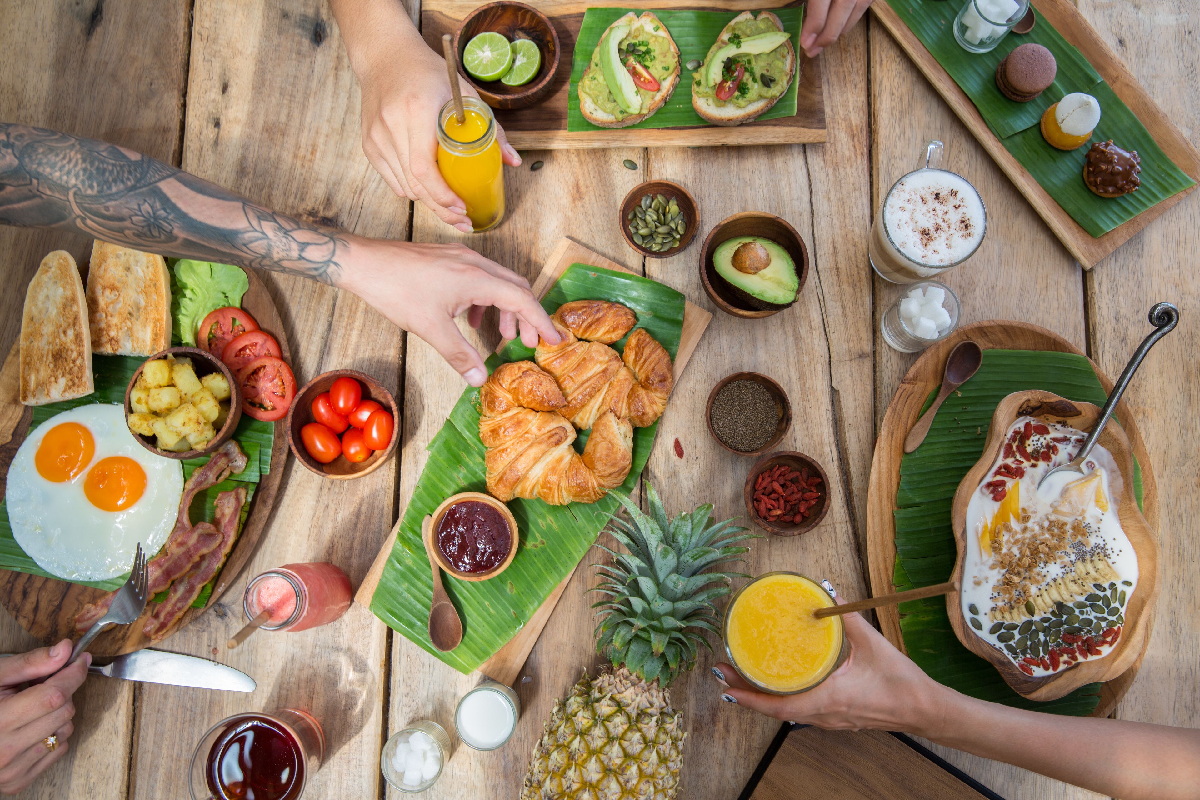 Ein Holztisch mit verschiedenen Frühstücksartikeln, darunter Eier, Toast, Croissants und tropische Früchte.