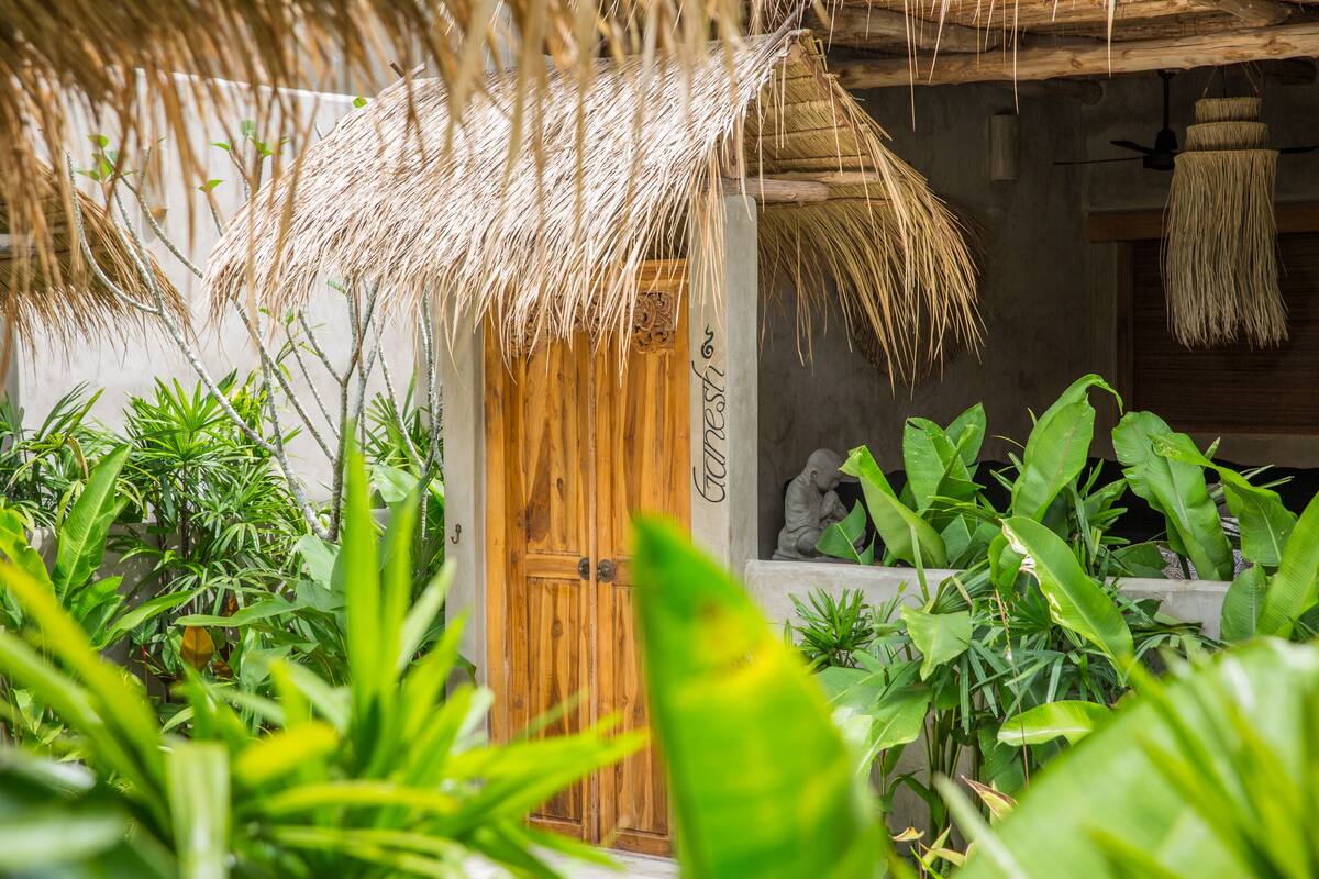 A wooden door with a thatched roof entrance, surrounded by lush green plants.