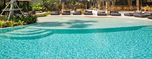 A clear blue pool with lounge chairs and umbrellas, surrounded by greenery and a bar area.