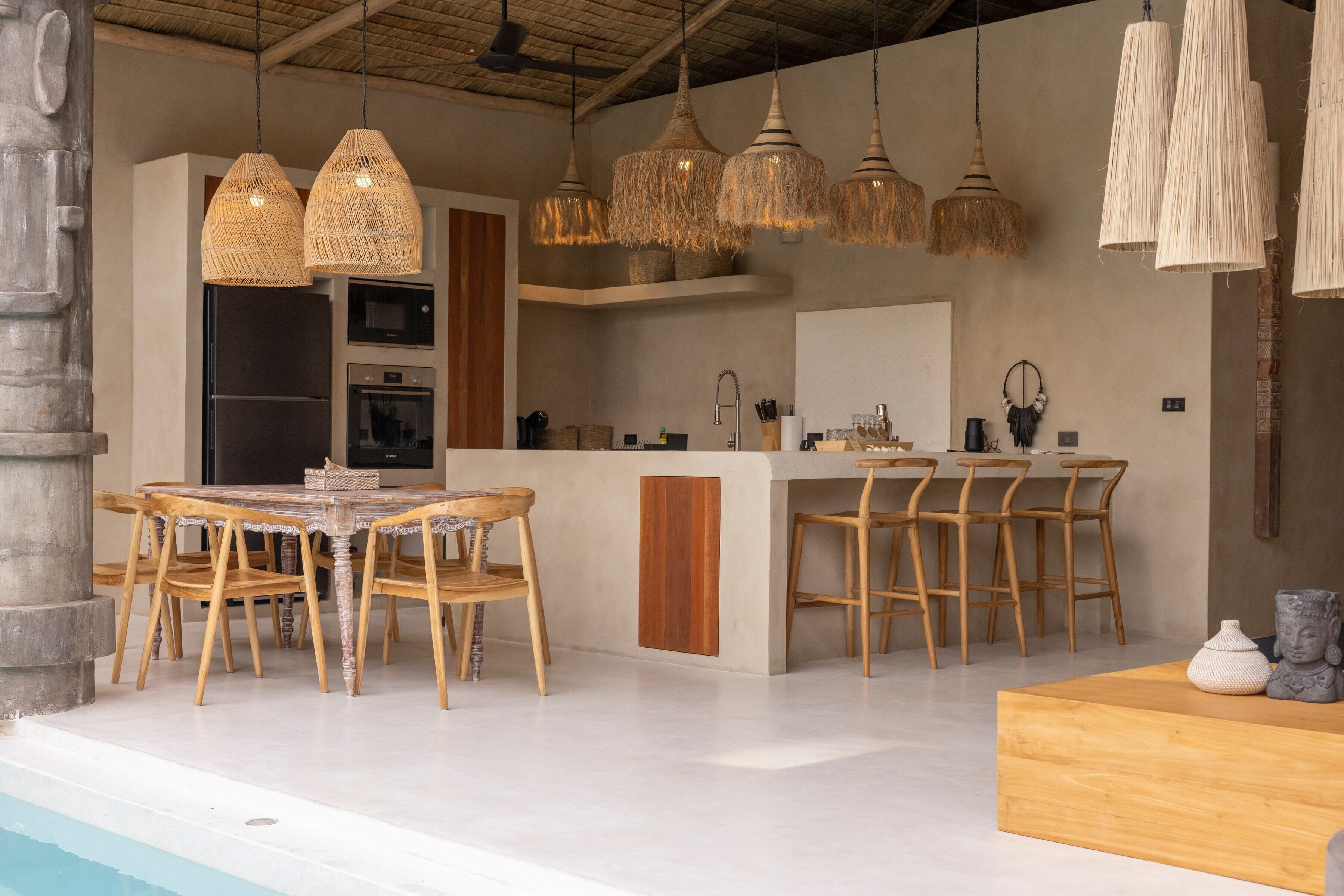 A modern kitchen with a central island, wooden chairs, hanging rattan lights, and a large window.