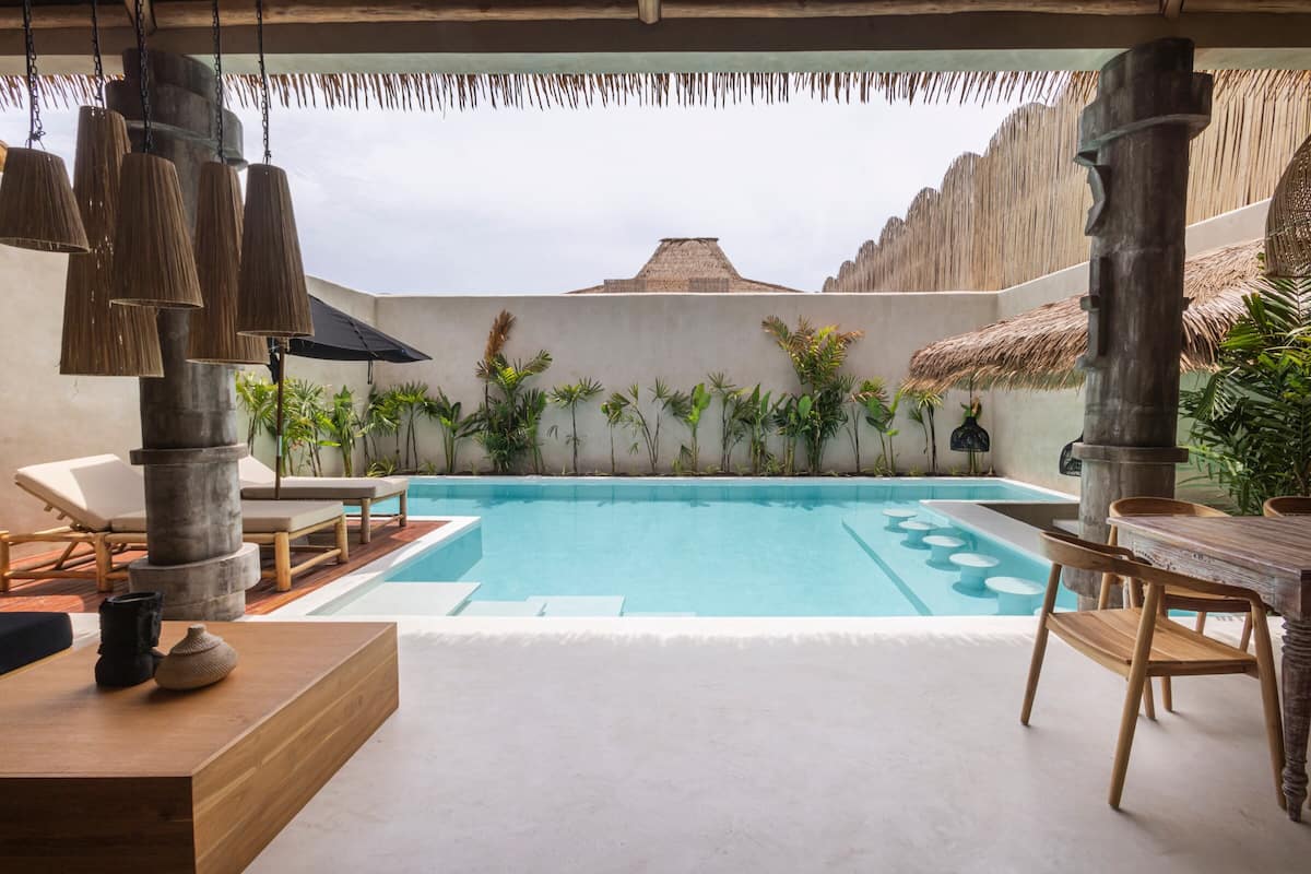 A pool area with lounge chairs, a wooden table, and hanging pendant lights.